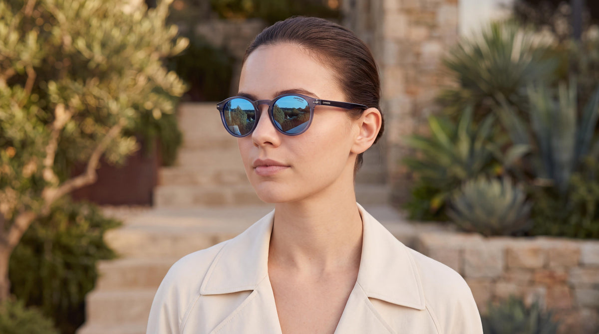 Refined woman in cream coat on a modern terrace, showcasing womens carrera blue round sunglasses with subtle blue tinted lenses in warm golden hour light.