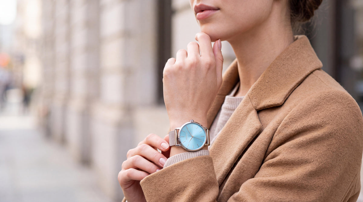 Woman in a camel coat touching her chin, wearing the Invicta Angel 48844 watch with an ice blue sunburst dial and silver mesh bracelet