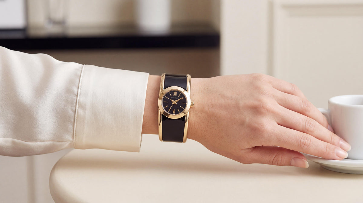 A woman's left hand rests on a cafe table, showcasing a womens caravelle black and gold bangle watch. Soft daylight illuminates the elegant scene.