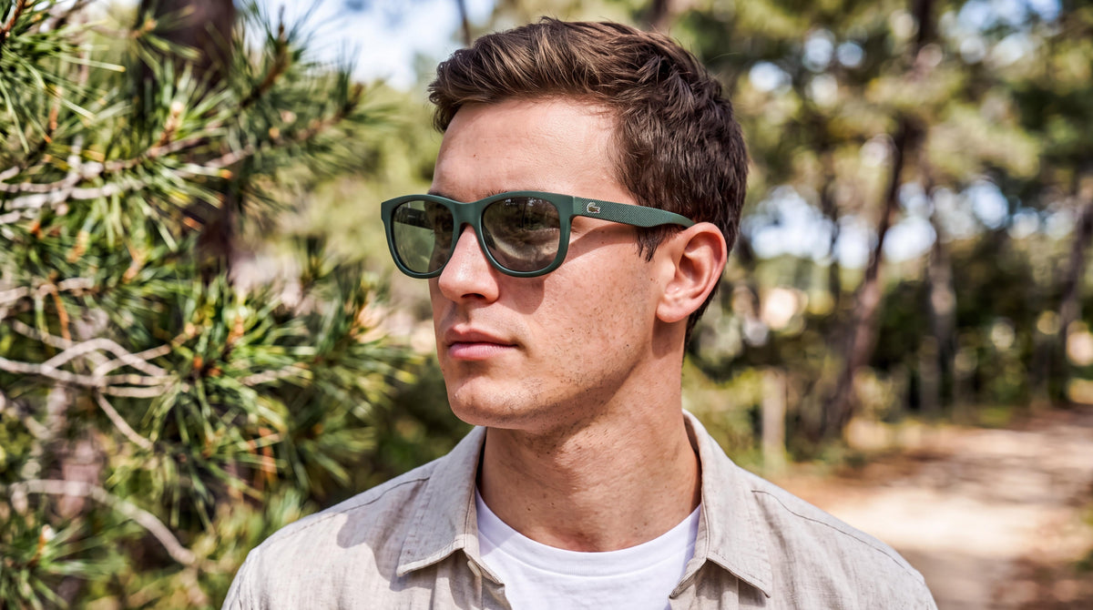 Man in a linen overshirt on a sunny pine path, wearing the Lacoste L848S matte dark green square sunglasses 