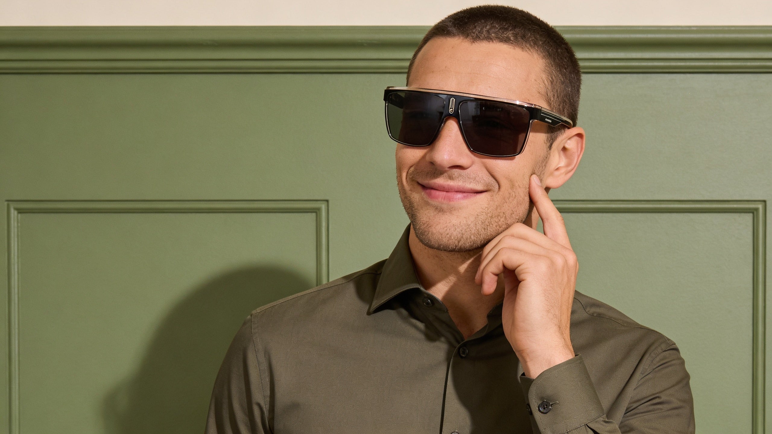 Fashion editorial half-body portrait featuring a subject wearing polarized sunglasses for outdoor activities, against a muted sage wall.