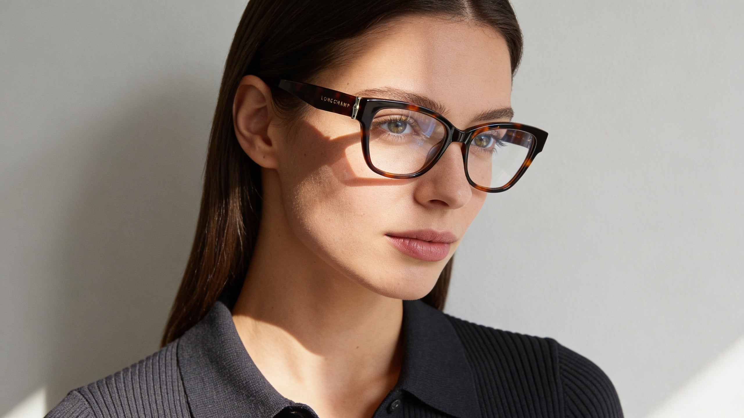 Refined editorial portrait of a woman wearing havana eyeglasses with gold accents, against a soft grey wall, calm expression.