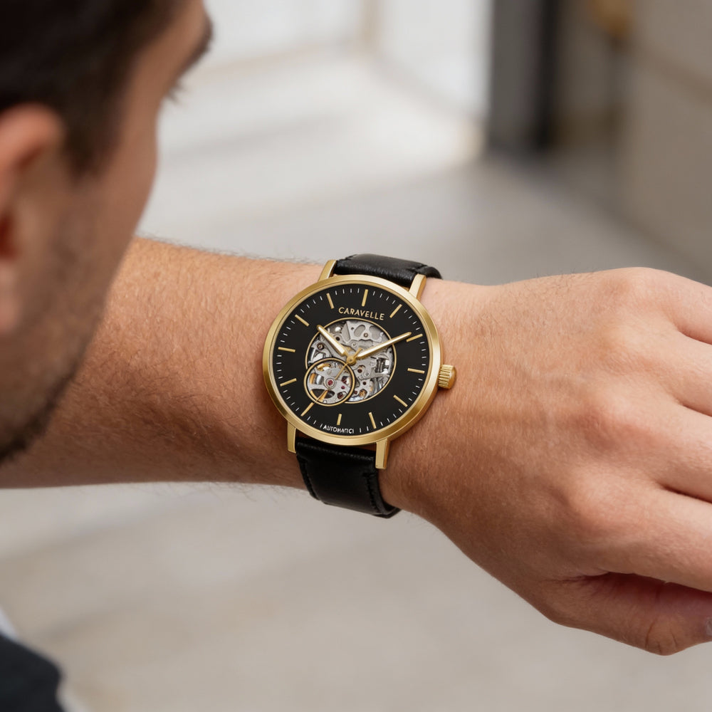 Reloj Caravelle clásico de vestir para hombre, automático, de acero inoxidable en tono dorado, con esqueleto y correa de piel negra.